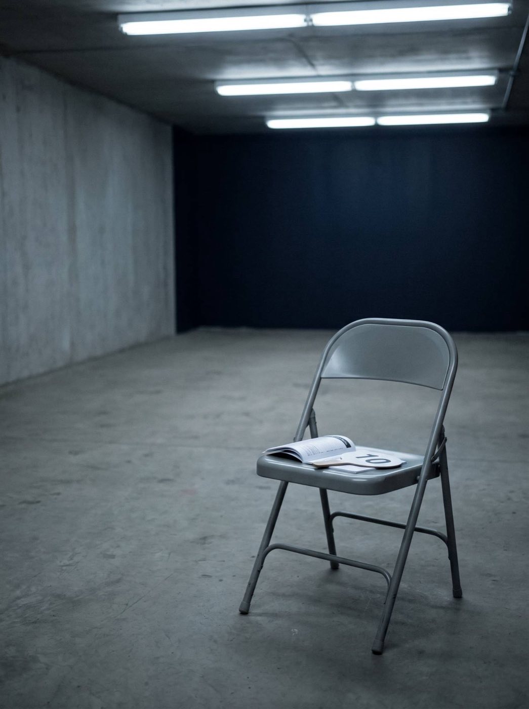 An abandoned bidder's card and auction brochure on an empty folding chair in a bare concrete room.